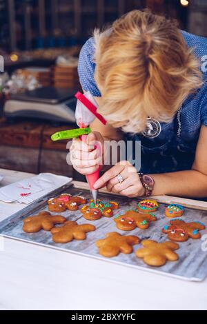 Unrecognizable women confectioner hand decorating a gingerman with a pastry bag, drawing a smile, making it cute, fun and delicious. Woman making ging Stock Photo