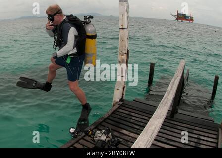 Man in scuba gear jumping into water from jetty with oil rig in background, Mabul Island, Sabah, Malaysia Stock Photo