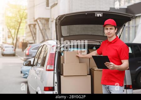 Express delivery by car. Deliveryman stands near open car and holds tablet, to check address Stock Photo