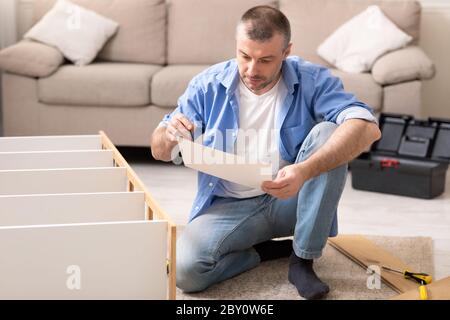 Man Assembling Cabinet Sitting Reading Installation Instruction At Home Stock Photo
