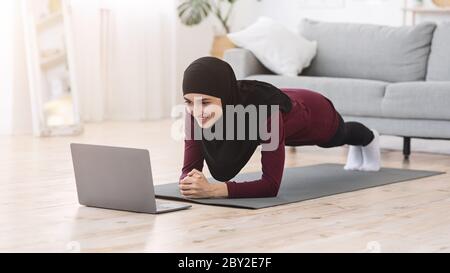 Home Trainings. Fit muslim woman doing yoga plank in front of laptop Stock Photo