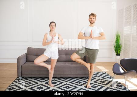 Young couple in white tshirts doing yoga at home standing in tree pose Stock Photo
