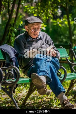 Bucharest/ Romania - 05.30.2020:Old man sitting on a bench in the Cismigiu park and reading a book. Stock Photo