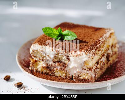 Perfect homemade tiramisu cake with fresh mint. Tiramisu portion on pink plate over white marble background. Delicious no bake tiramisu in natural daylight. Close up Stock Photo