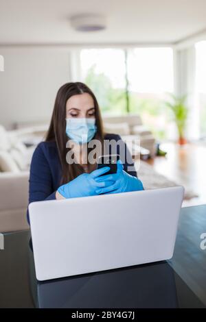 Young businesswoman with surgical mask and gloves using mobile phone and working online from home, covid-19 isolation concept. Stock Photo
