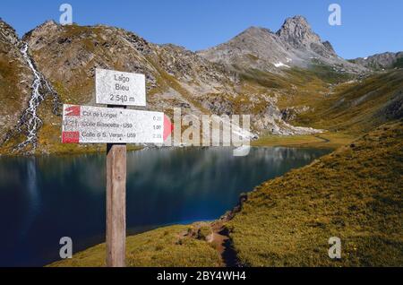 The Lac Bleu in Chianale, mountain lake in the italian alps of Cuneo, Piedmont, with sign for trekkers pointing in direction of nearby passes named co Stock Photo