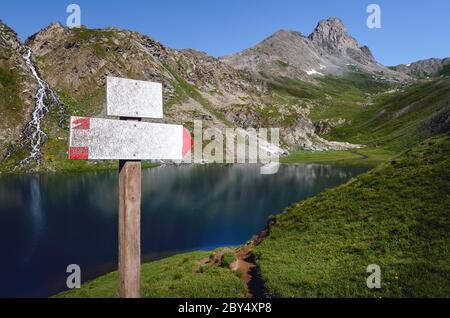 The Lac Bleu in Chianale, mountain lake in the italian alps of Cuneo, Piedmont, with white blank sign for trekkers pointing at the nearby peak Stock Photo