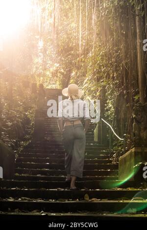 Rear view of beautiful fashionable stylish female traveler wearing a hat, walking the staircase of traditional hidu tample around Ubud on Bali Stock Photo