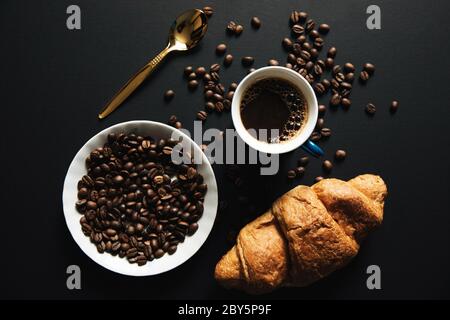 Coffee mug with roasted coffee beans,crossiant and golden spoon on dark matte finish wooden table. View from top Stock Photo