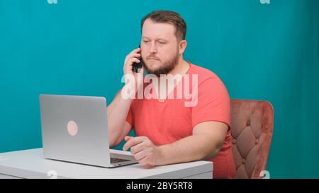 Young happy businessman in a pink t-shirt is working on a laptop and talking on a cell phone on a green screen. Portrait of a male talking man looking Stock Photo