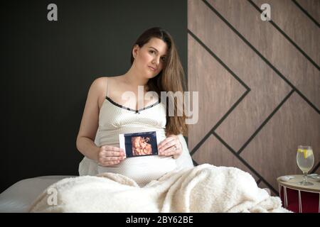 Pregnant woman holding ultrasound image. Concept of pregnancy, health care, gynecology, medicine. Young mother waiting of the baby. Stock Photo