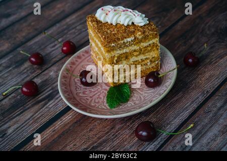 Piece of multilayer honey cake with cream. Classic honey cake pieces with custard. Homemade honey cake on wooden background with cherry and mint leaves. Delicious slice of layered tart on plate Stock Photo