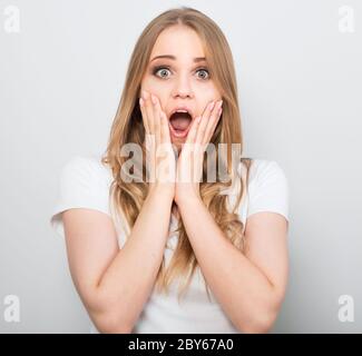 Pretty excited woman with fair blond hair wide opening mouth and big eyes in casual dress and holding hands near the mouth. Studio shot of beautiful w Stock Photo