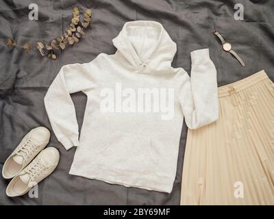 Fashionable female look with white empty hoody, cream pleated skirt and white sneakers. Top view of white blank hoody with long sleeves over gray bed linen. Mock up for hoody print design. Stock Photo