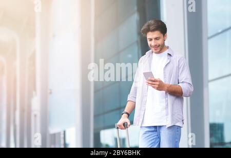 Ordering Taxi. Man Using Smartphone While Standing With Suitcase Near Airport Stock Photo