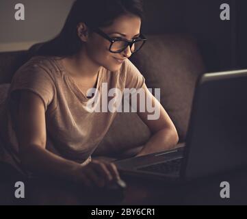 Smiling thinking young beautiful woman in eye glasses looking on computer monitor and working on dark shadow home background. Toned vintage closeup po Stock Photo