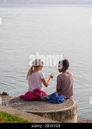 Two little girls, one blonde and the other black, sit on the edge of a lake in Italy and talk to each other... Stock Photo