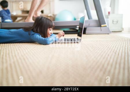 Girl using laptop on living room floor next to father on treadmill Stock Photo
