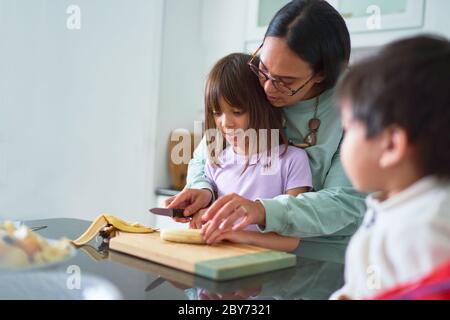 Mother helping daughter cut banana in kitchen Stock Photo