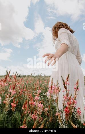 Back view on an angle of young woman in white long dress walking with hand out in a blossoming flower field Stock Photo