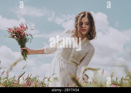 Upper body portrait of young woman in long white dress walking with a flower bouquet leaning to one side, against blue sky with clouds Stock Photo