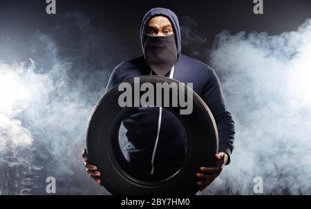 Photo of astonished afro american guy hold car vehicle transport tyre wear jumper sweater bandanna isolated over smoke black color background Stock Photo