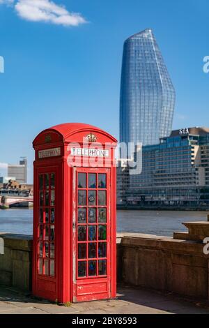 Traditional red London public telephone box on the embankment in London, England overlooking the River Thames with skyscrapers in the background Stock Photo