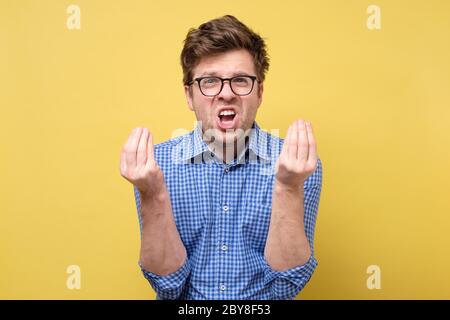 Young handsome man showing italian gesture that means what do you want over yellow background. He has a quarrel with his boss or coworker. Studio shot Stock Photo