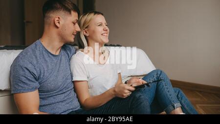 Side view photo of a caucasian woman with blonde hair laughing on the floor with her lover using a tablet Stock Photo