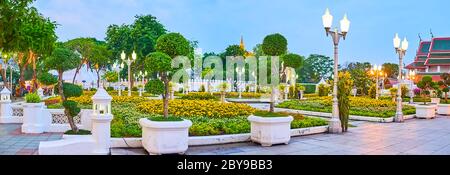 The evening panorama of ornamental Mahajetsadabadin park with topiary trees in pots, marigold flower beds, vintage lanterns and hanging orchids, Bangk Stock Photo