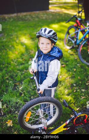 Caucasian child boy in a helmet learns to repair his bike. Child cyclist checks the mechanism of a bicycle in a clearing of green grass in the park Stock Photo