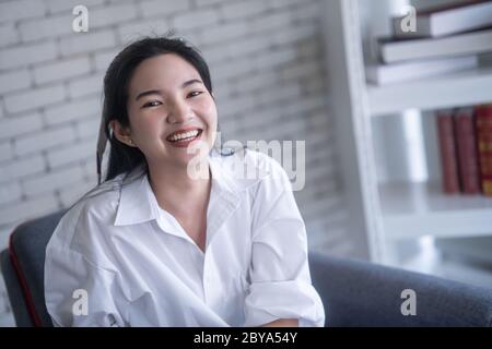 beautiful young asian woman smiling while sitting on the chair against white brick wall Stock Photo