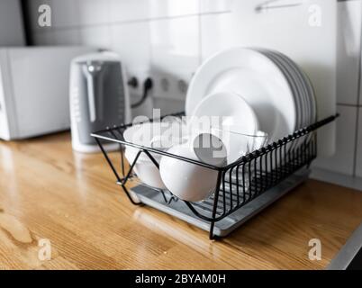 Dish rack holds many dishes and cups against wooden countertop, white wall tiles, sink and faucet. Budget and lightweight antimicrobial dish drainer Stock Photo