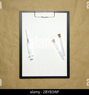 The tablet with a clip for the clip of paper with a white sheet a4 lies against the background of craft brown crumpled paper. On top is a medical syringe and two transparent empty tubes with stoppers. Place for text and layout for design. Stock Photo