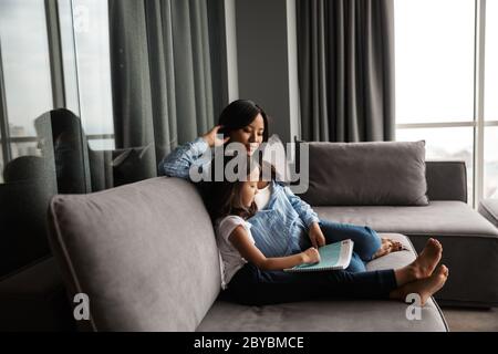 Photo of pleased pregnant asian woman with her little daughter reading book while sitting on couch at home Stock Photo