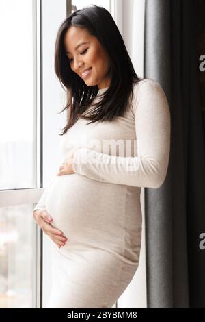 Photo of happy pregnant asian woman smiling and touching her belly while standing near window Stock Photo