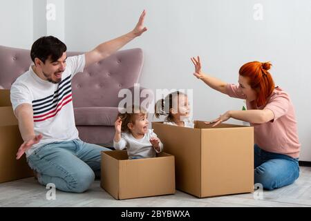 Funny active family playing on moving day, excited happy adult parents mom dad playing with cardboard boxes with cute little kids sit inside having fu Stock Photo