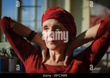 Female portrait of mature woman dressed in red clothes and sitting at the end of her work day on sewing machine and cotton bobbins background, tailor Stock Photo