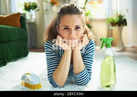 Portrait of modern woman in striped t-shirt and white pants with spray bottle of green cleaning supplies and yellow brush laying on white carpet at mo Stock Photo
