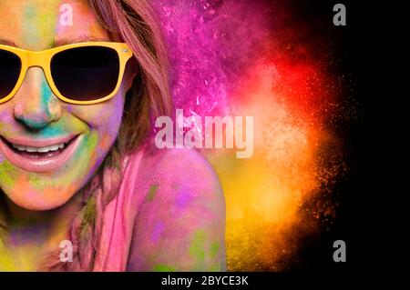 Happy young woman wearing trendy yellow sunglasses enjoying celebrating Holi fest covered in vibrant colorful powder pink, magenta and yellow with bur Stock Photo