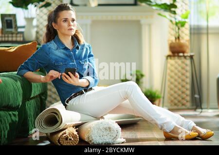 pensive modern middle age woman in jeans shirt and white pants with pile of carpets searching for carpet cleaning service on a smartphone at modern ho Stock Photo