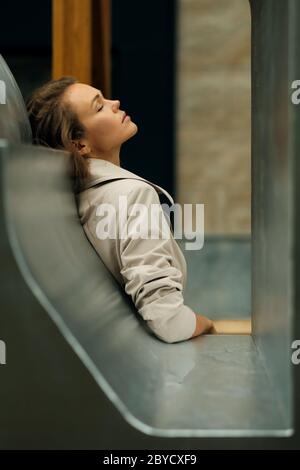 Attractive young lady wearing gray trench coat lean on architectural element in downtown with eyes closed to relax. Stock Photo
