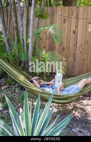 A mother and her little boy lie in a hammock and read a picture book together surrounded by greenery in their backyard in Miami, Florida. Stock Photo