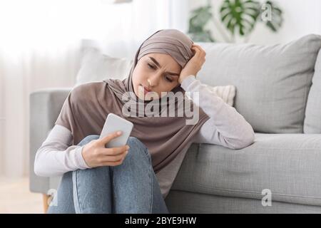Depression Concept. Upset Arabic Woman Sitting With Smartphone On Floor At Home