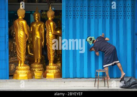 An employee of a shop in Bamrung Muang Rd., Bangkok, Thailand, cleaning the door shutters, Buddha statues on sale seen behind the half-opened shutter Stock Photo