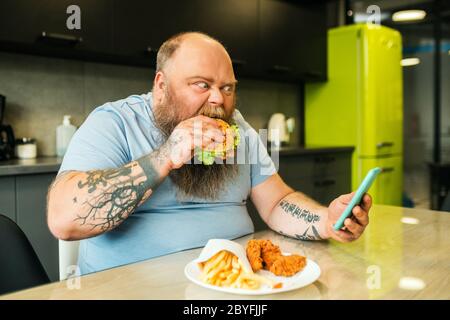 Bearded fat man looking at smartphone biting burger Stock Photo