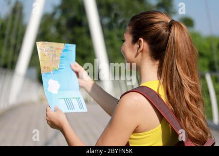 Back view of young female backpacker looking at a map on bridge in the forest. Girl reading a map for her journey in nature early morning on summer. Stock Photo