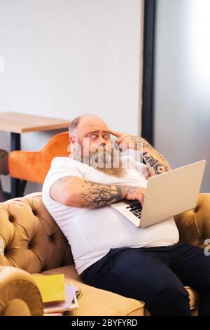 Bald bearded plump man sitting with a laptop in hands and looking thoughtful Stock Photo