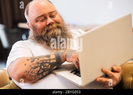 Bald bearded plump man sitting with a laptop in hands and smiling Stock Photo