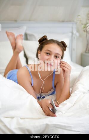 Young girl listening to music with earphones. Girl lies in bed in the morning. Free time outside of school. Quarantine life. Stock Photo